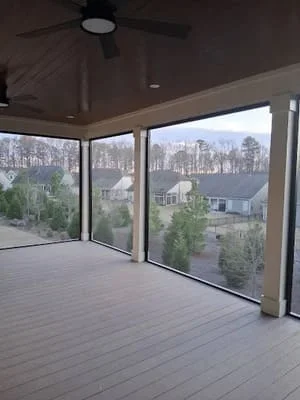 Elevated screen room with composite deck, glass panels, and ceiling fan overlooking wooded neighborhood